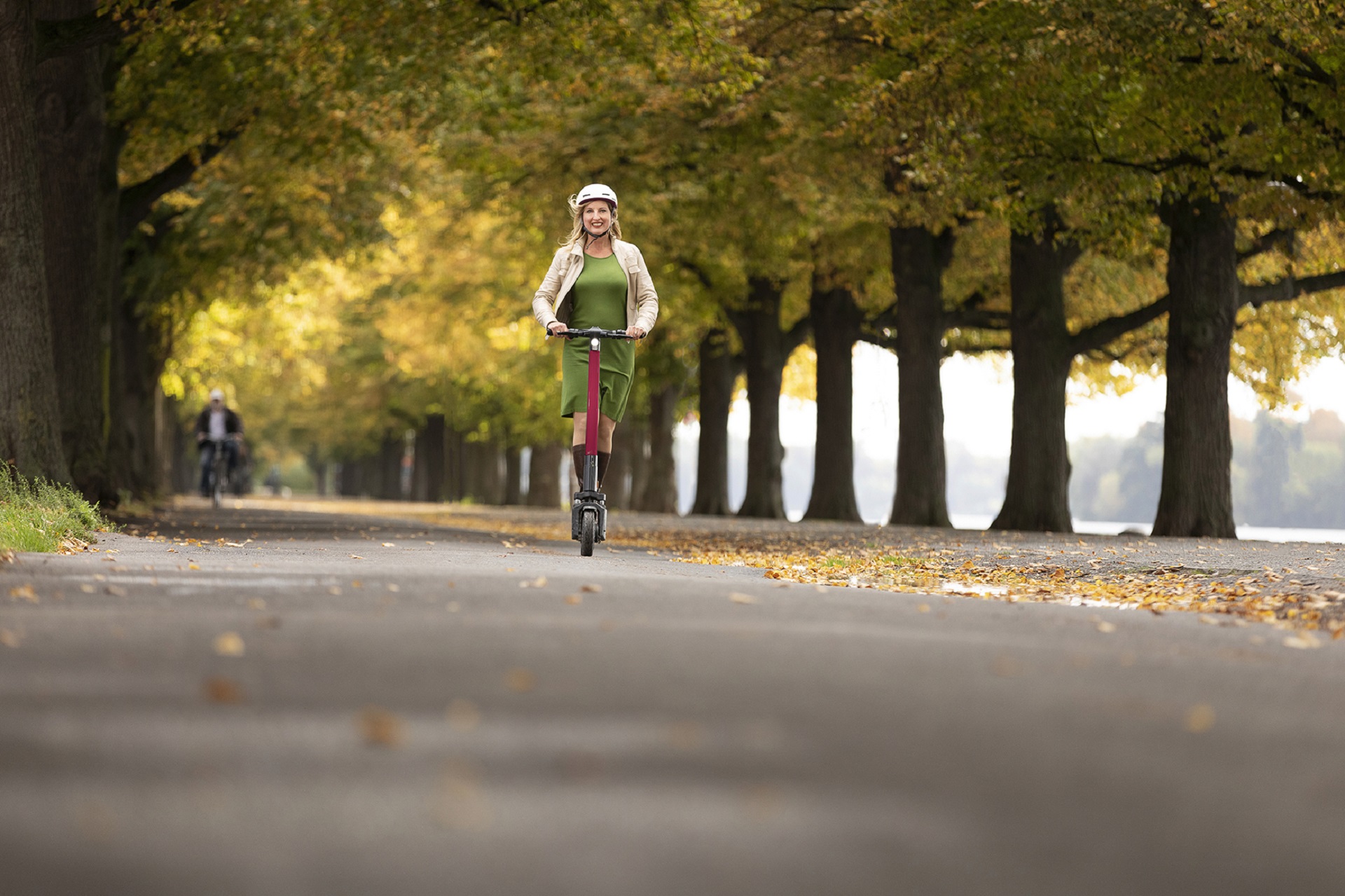 Frau faährt einen E-Scooter auf einem von Bäumen gesäumten Fahrradweg.