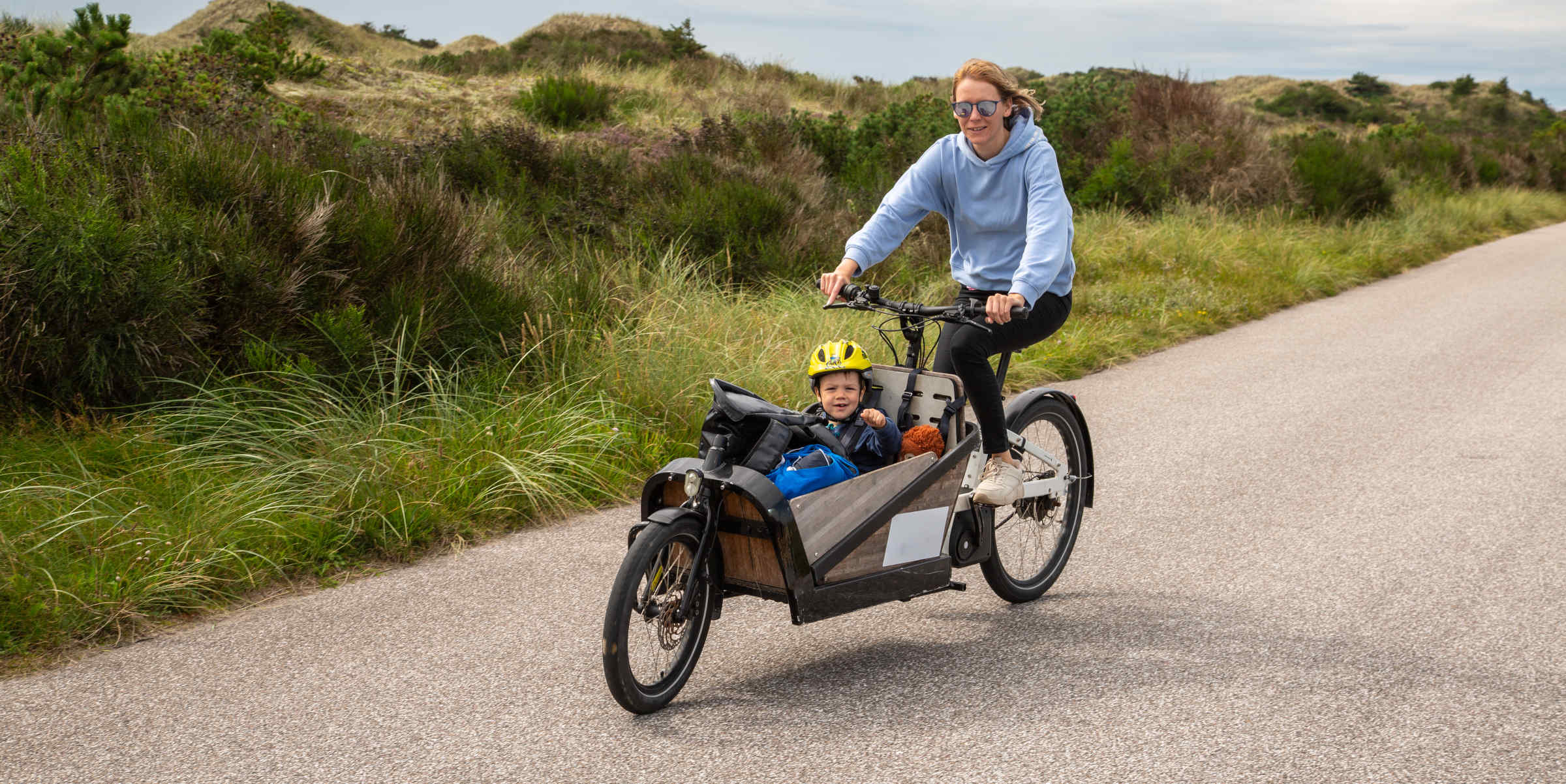 Frau fährt mit Kind im Lastenrad durch Dünen.