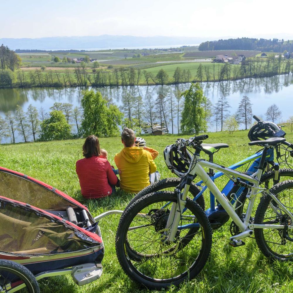 Zwei Personen sitzen am See-Ufer. Im Vordergrund befinden sich zwei Fahrränder. Eines davon hat einen Anhänger.