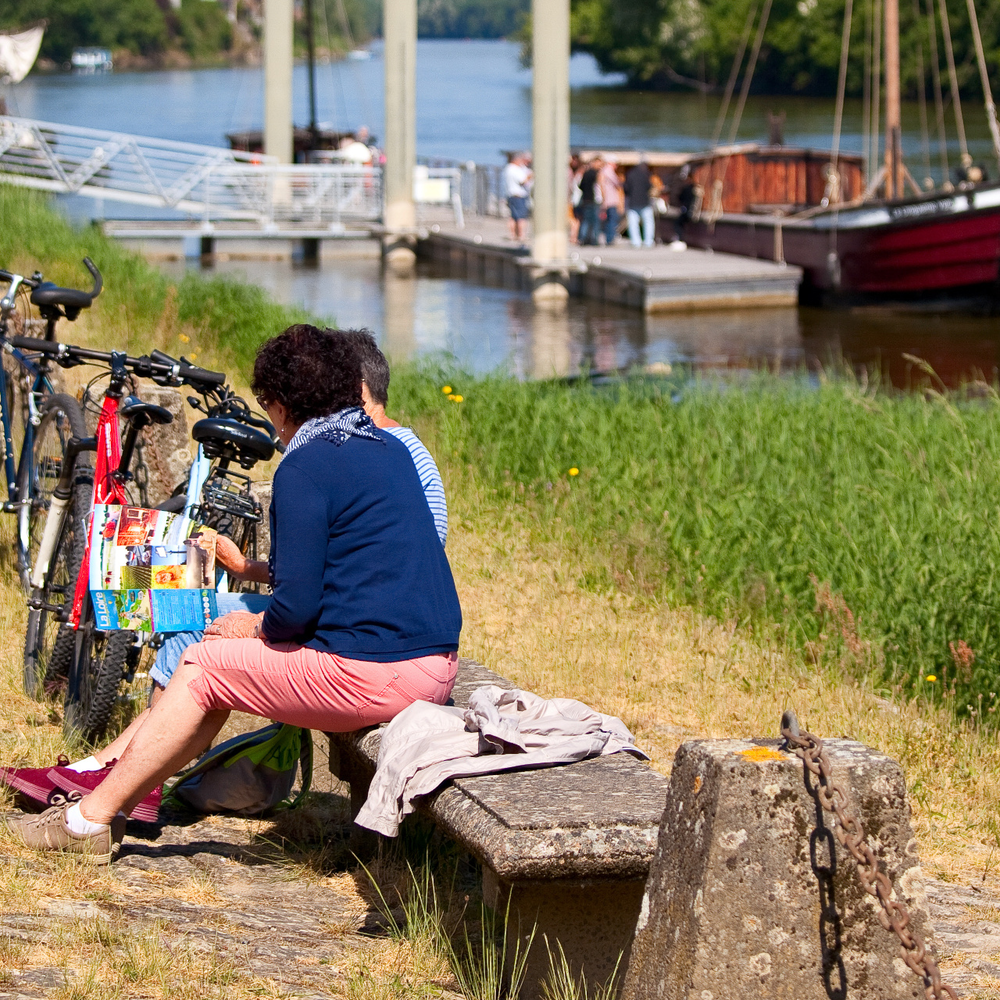 Zwei Personen sitzen auf einer Bank am Rande eines Radweges und schauen in ein Prospekt. Neben Ihnen stehen meherere Fahrräder. Am Radweg führt ein Fluss vorbei. Im Hintergrund ist ein Boot zu sehen.